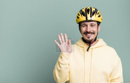 Photo for Adult man smiling happily, waving hand, welcoming and greeting you. bike helmet and bicycle concept - Royalty Free Image