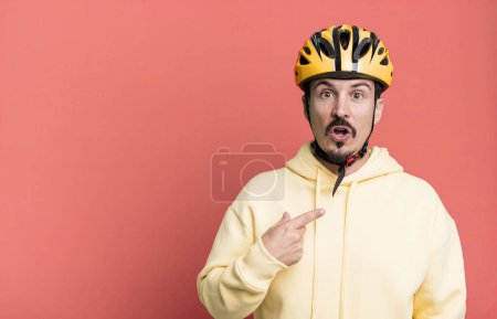 Foto de Adult man looking shocked and surprised with mouth wide open, pointing to self. bike helmet and bicycle concept - Imagen libre de derechos