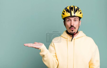 Foto de Adult man looking surprised and shocked, with jaw dropped holding an object. bike helmet and bicycle concept - Imagen libre de derechos