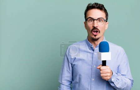Téléchargez les photos : Adult man looking very shocked or surprised with a microphone. presenter or journalist concept - en image libre de droit