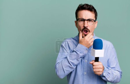 Foto de Adult man with mouth and eyes wide open and hand on chin with a microphone. presenter or journalist concept - Imagen libre de derechos