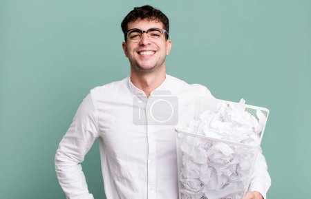 Téléchargez les photos : Homme adulte souriant heureux avec une main sur la hanche et confiant. boules de papier erreurs poubelle - en image libre de droit