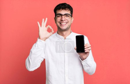 Téléchargez les photos : Homme adulte se sentant heureux, montrant l'approbation avec le geste correct et montrant son écran de smartphone - en image libre de droit