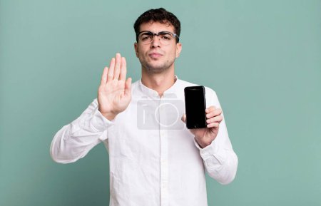 Téléchargez les photos : Homme adulte regardant sérieux montrant la paume ouverte faisant geste d'arrêt et montrant son écran de smartphone - en image libre de droit