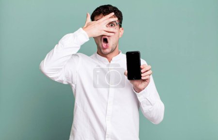 Téléchargez les photos : Homme adulte regardant choqué, effrayé ou terrifié, couvrant le visage avec la main et montrant son écran de smartphone - en image libre de droit