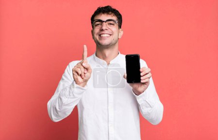 Téléchargez les photos : Homme adulte souriant fièrement et en toute confiance faisant numéro un et montrant son écran de smartphone - en image libre de droit