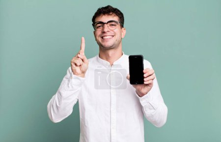 Foto de Hombre adulto sonriendo y buscando amigable, mostrando el número uno y mostrando la pantalla de su teléfono inteligente - Imagen libre de derechos