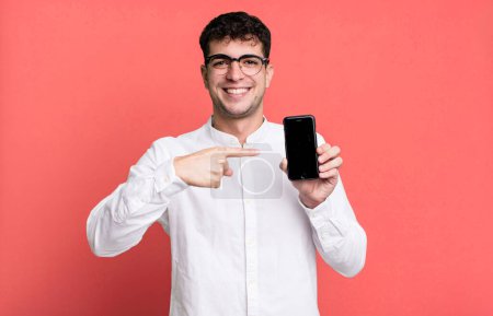 Téléchargez les photos : Homme adulte souriant joyeusement, se sentant heureux et pointant vers le côté et montrant son écran de smartphone - en image libre de droit