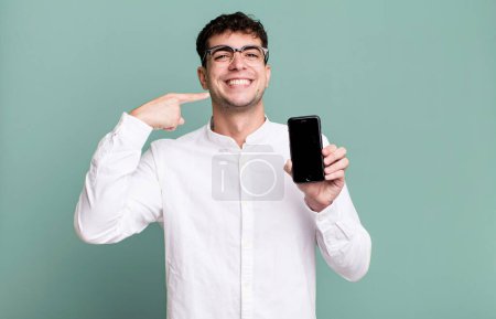 Téléchargez les photos : Homme adulte souriant avec confiance pointant vers son propre sourire large et montrant son écran de smartphone - en image libre de droit