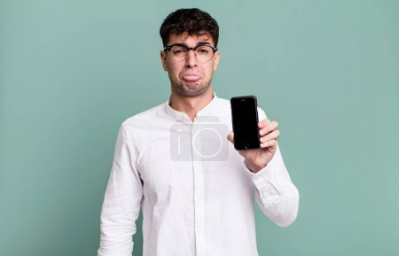 Téléchargez les photos : Homme adulte se sentant triste et pleurnichard avec un regard malheureux et pleurer et montrant son écran de smartphone - en image libre de droit