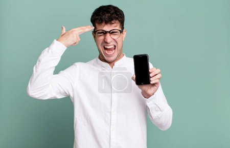 Téléchargez les photos : Homme adulte regardant malheureux et stressé, geste de suicide faisant signe d'arme à feu et montrant son écran de smartphone - en image libre de droit