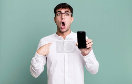 Téléchargez les photos : Homme adulte regardant choqué et surpris avec la bouche grande ouverte, pointant vers soi-même et montrant son écran smartphone - en image libre de droit