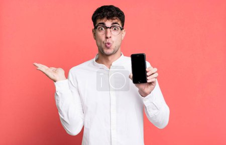 Téléchargez les photos : Homme adulte regardant surpris et choqué, avec la mâchoire tombée tenant un objet et montrant son écran de smartphone - en image libre de droit