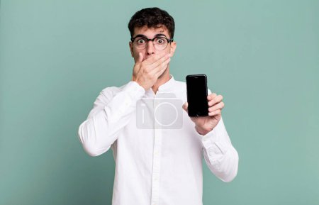 Téléchargez les photos : Homme adulte couvrant la bouche avec les mains avec un choc et montrant son écran de smartphone - en image libre de droit
