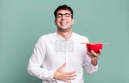 Foto de Hombre adulto riendo en voz alta en algún chiste hilarante sosteniendo un cuenco de fideos ramen - Imagen libre de derechos