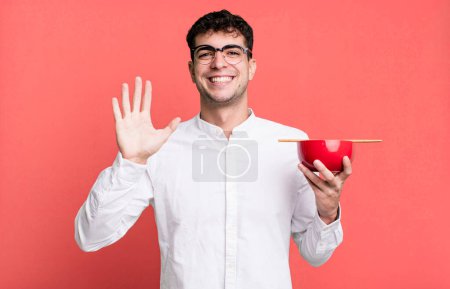 Foto de Hombre adulto sonriendo felizmente, saludando con la mano, dándote la bienvenida y saludándote sosteniendo un tazón de fideos ramen - Imagen libre de derechos