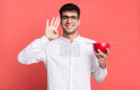 Foto de Hombre adulto sonriendo y buscando amigable, mostrando el número cuatro sosteniendo un tazón de fideos ramen - Imagen libre de derechos