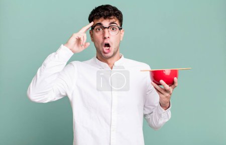 Téléchargez les photos : Homme adulte regardant surpris, réalisant une nouvelle pensée, idée ou concept tenant un bol de nouilles ramen - en image libre de droit