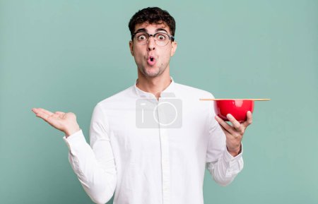 Photo for Adult man looking surprised and shocked, with jaw dropped holding an object holding a ramen noodles bowl - Royalty Free Image