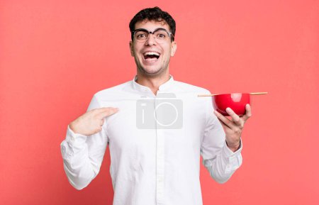 Foto de Hombre adulto mirando sorprendido y sorprendido con la boca abierta, apuntando a uno mismo sosteniendo un tazón de fideos ramen - Imagen libre de derechos