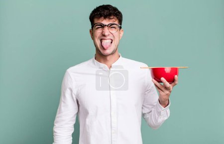 Téléchargez les photos : Homme adulte se sentant dégoûté et irrité et la langue tenant un bol de nouilles ramen - en image libre de droit