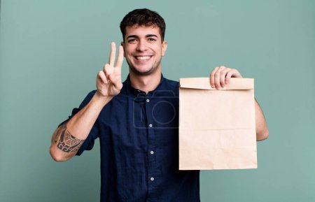 Photo for Adult man smiling and looking happy, gesturing victory or peace with a take away breakfast paper bag with a take away breakfast paper bag - Royalty Free Image