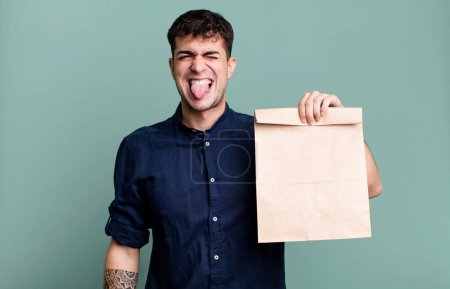 Photo for Adult man with cheerful and rebellious attitude, joking and sticking tongue out with a take away breakfast paper bag with a take away breakfast paper bag - Royalty Free Image