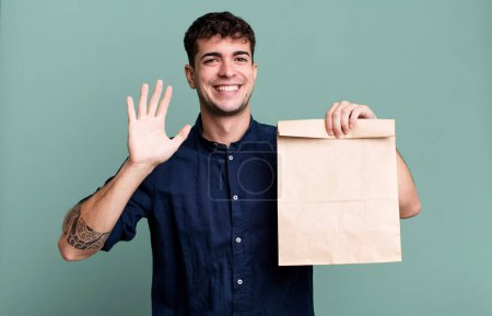 Photo for Adult man smiling happily, waving hand, welcoming and greeting you with a take away breakfast paper bag with a take away breakfast paper bag - Royalty Free Image