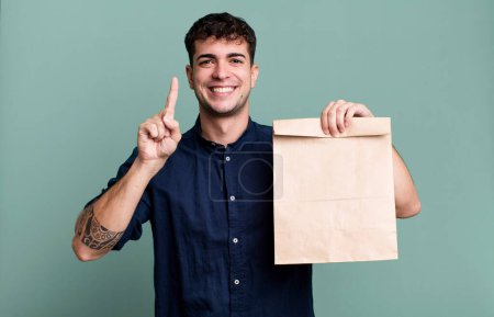 Téléchargez les photos : Homme adulte souriant et regardant amical, montrant numéro un avec un sac à emporter petit déjeuner en papier avec un sac à emporter petit déjeuner en papier - en image libre de droit