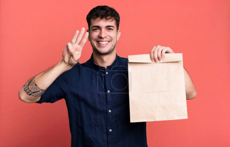 Téléchargez les photos : Homme adulte souriant et regardant amical, montrant numéro trois avec un sac à emporter petit déjeuner en papier avec un sac à emporter petit déjeuner en papier - en image libre de droit