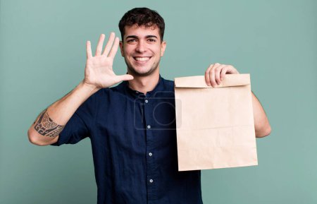 Foto de Hombre adulto sonriendo y buscando amigable, mostrando el número cinco con una bolsa de papel para llevar con una bolsa de papel para llevar - Imagen libre de derechos