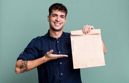 Téléchargez les photos : Homme adulte souriant joyeusement, se sentant heureux et montrant un concept avec un sac en papier petit déjeuner à emporter avec un sac en papier petit déjeuner à emporter - en image libre de droit