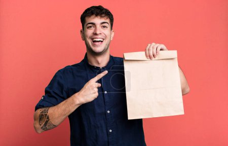 Téléchargez les photos : Homme adulte regardant excité et surpris pointant vers le côté avec un sac en papier à emporter petit déjeuner avec un sac en papier à emporter petit déjeuner - en image libre de droit