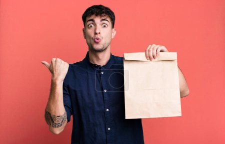 Téléchargez les photos : Homme adulte regardant étonné dans l'incrédulité avec un sac de papier de petit déjeuner à emporter avec un sac de papier de petit déjeuner à emporter - en image libre de droit