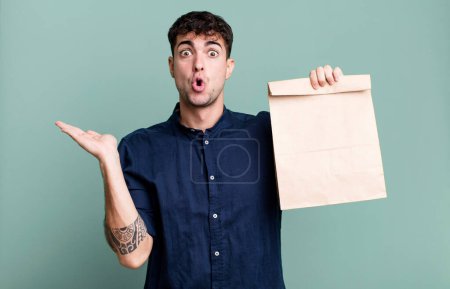 Foto de Hombre adulto mirando sorprendido y sorprendido, con la mandíbula caída sosteniendo un objeto con una bolsa de papel para llevar con una bolsa de papel de desayuno para llevar - Imagen libre de derechos