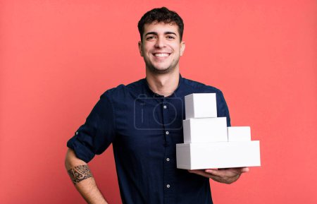 Téléchargez les photos : Homme adulte souriant heureux avec une main sur la hanche et confiant avec des emballages de produits vierges - en image libre de droit