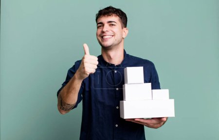Téléchargez les photos : Homme adulte se sentant fier, souriant positivement avec les pouces vers le haut avec des emballages de produits vierges - en image libre de droit