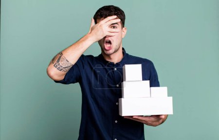 Foto de Hombre adulto mirando sorprendido, asustado o aterrorizado, cubriendo la cara con la mano con paquetes de productos en blanco - Imagen libre de derechos