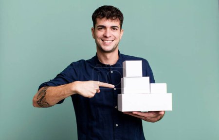 Foto de Hombre adulto sonriendo alegremente, sintiéndose feliz y señalando hacia un lado con paquetes de productos en blanco - Imagen libre de derechos