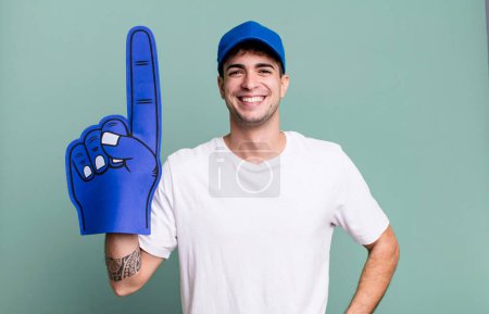 Foto de Hombre adulto sonriendo felizmente con una mano en la cadera y confiado. concepto de ventilador número uno - Imagen libre de derechos