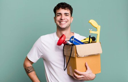 Foto de Hombre adulto sonriendo felizmente con una mano en la cadera y seguro con una caja de herramientas. concepto de ama de llaves - Imagen libre de derechos