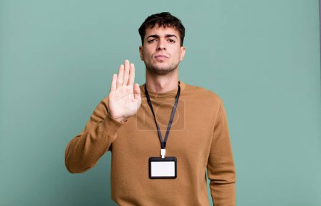 Foto de Hombre adulto mirando serio mostrando palmera abierta haciendo gesto de parada con una tarjeta de identidad de acceso - Imagen libre de derechos