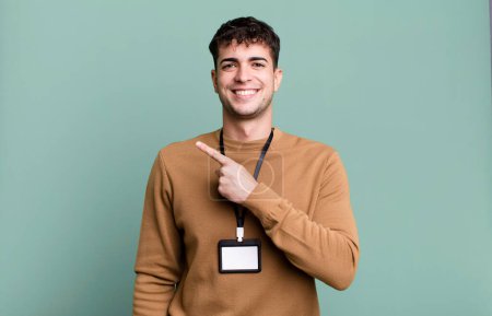 Foto de Hombre adulto sonriendo alegremente, sintiéndose feliz y señalando a un lado con una tarjeta de identidad de acceso - Imagen libre de derechos