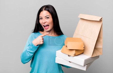 Foto de Hispanic pretty woman looking excited and surprised pointing to the side. with take away fast food packages - Imagen libre de derechos