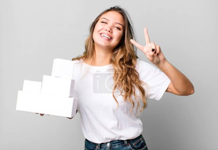 Photo for Hispanic pretty woman smiling and looking friendly, showing number two with white boxes packages - Royalty Free Image