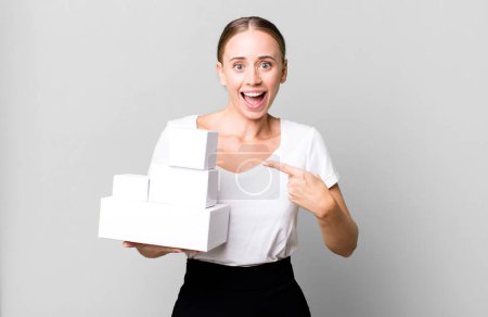 Photo for Caucasian pretty woman looking excited and surprised pointing to the side with white boxes packages - Royalty Free Image