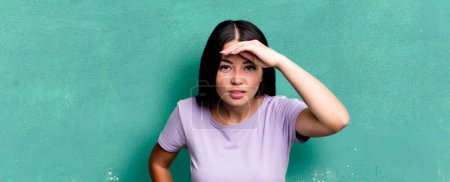 Photo for Pretty latin woman looking bewildered and astonished, with hand over forehead looking far away, watching or searching - Royalty Free Image
