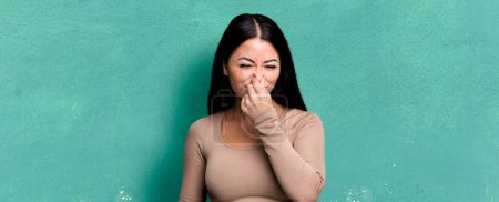 Téléchargez les photos : Jolie femme latine se sentant dégoûtée, tenant le nez pour éviter de sentir une puanteur sale et désagréable - en image libre de droit