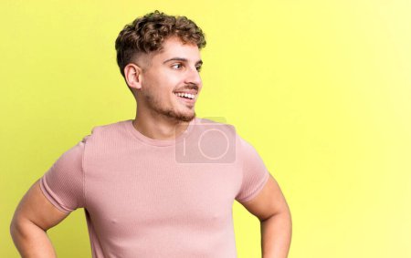 Photo for Young adult caucasian man looking happy, cheerful and confident, smiling proudly and looking to side with both hands on hips - Royalty Free Image