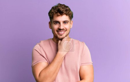 Téléchargez les photos : Young adult caucasian man looking happy and smiling with hand on chin, wondering or asking a question, comparing options - en image libre de droit